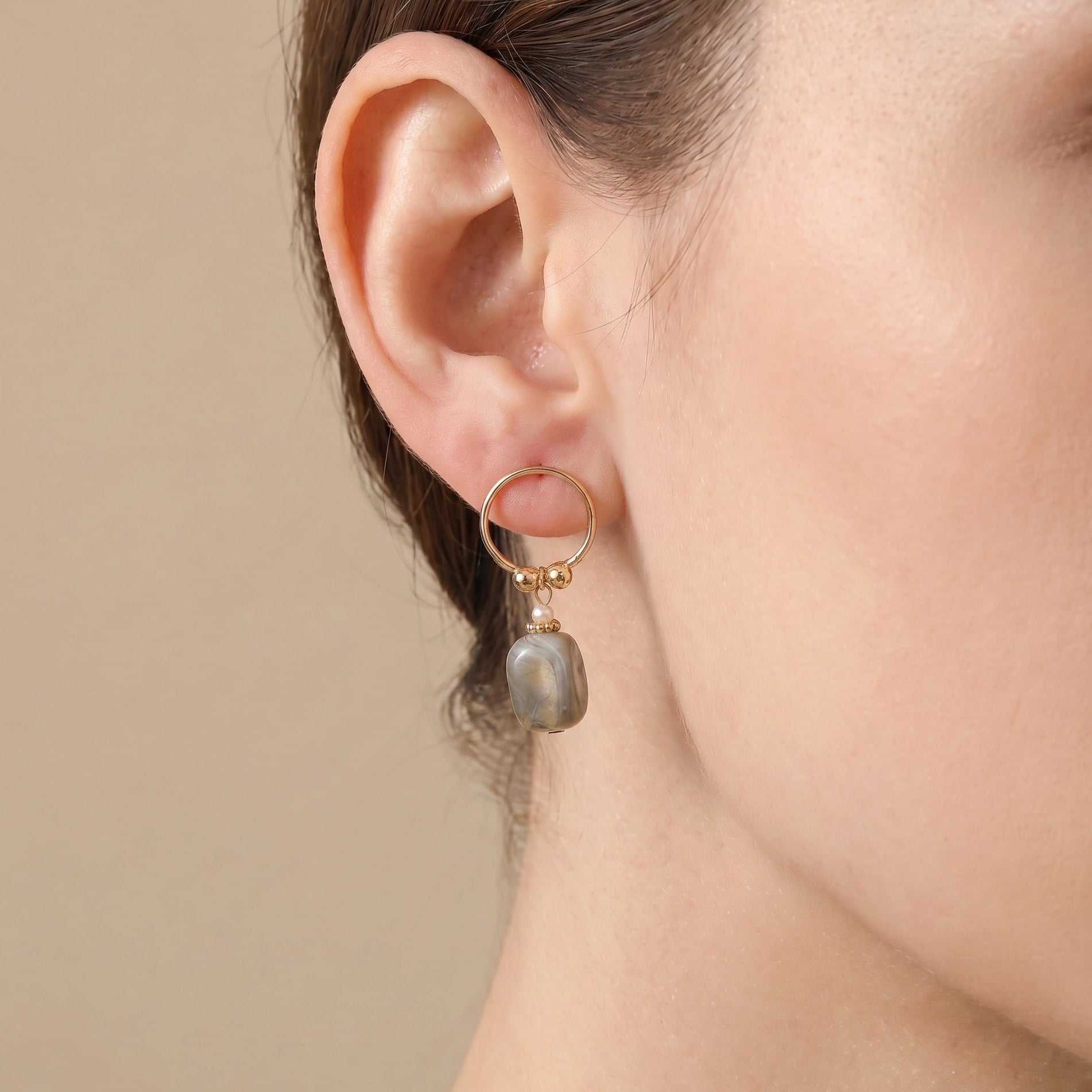 Close-up of an ear wearing a gold hoop earring with a gray stone pendant against a beige background